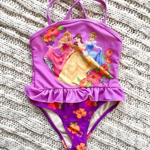 Puede incluir: Un traje de ba&ntilde;o morado con un dise&ntilde;o que presenta tres princesas animadas y detalles florales. La parte superior tiene un detalle de volantes y tirantes finos. La parte inferior es morada oscura con flores naranjas y rosas. Para ni&ntilde;os.