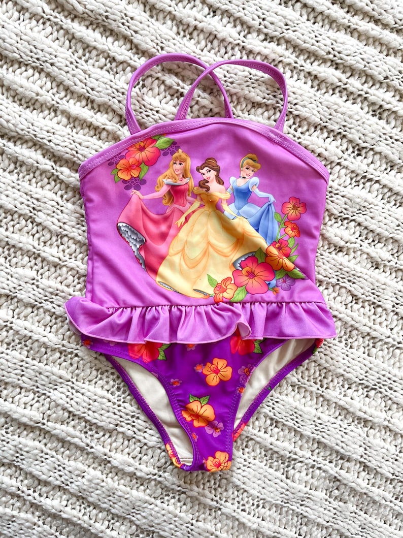 Puede incluir: Un traje de ba&ntilde;o morado con un dise&ntilde;o que presenta tres princesas animadas y detalles florales. La parte superior tiene un detalle de volantes y tirantes finos. La parte inferior es morada oscura con flores naranjas y rosas. Para ni&ntilde;os.