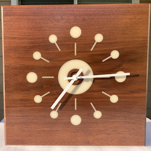 May include: A modern, minimalist clock with a white face and white hands. The clock has a wood frame and a unique design with white circles and lines.