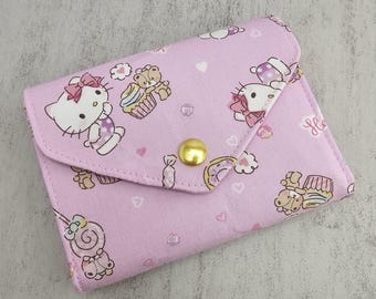 Puede incluir: Una cartera de tela rosa con un patrón repetido de Hello Kitty, cupcakes y otras ilustraciones lindas. La cartera tiene un cierre de solapa asegurado por un broche dorado. El diseño general es caprichoso y juguetón.