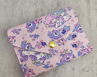 Puede incluir: Una cartera de tela rosa con un estampado floral y un cierre de botón a presión dorado. La cartera presenta un patrón repetido de flores azules, moradas y rosas, y una ilustración de dibujos animados de dos personajes. La cartera está cerrada.