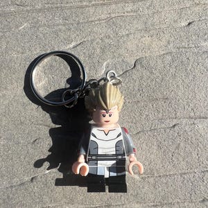 Pode incluir: Um chaveiro Lego com uma minifigura com penteado loiro, vestindo uma roupa cinza e branca com calças pretas. O chaveiro tem um anel prateado para prender chaves.