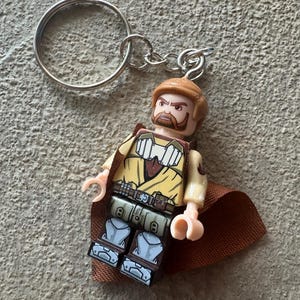 Op de afbeelding: Een Lego-stijl sleutelhanger met een personage met een bruine baard, haar en een beige en bruine outfit. De figuur heeft een bruine cape en is bevestigd aan een zilveren metalen sleutelhanger.