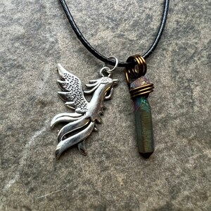 May include: A black cord necklace featuring two pendants. One is a silver phoenix charm, and the other is a dark, iridescent crystal wrapped in copper wire. The necklace rests on a textured, gray surface.