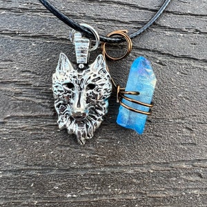 Pode incluir: Um colar com um pingente de cabeça de lobo prateado e um amuleto de cristal azul em um cordão preto.