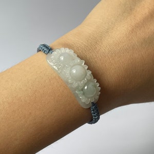 Op de afbeelding: Een jade armband met een gesneden zonnebloemontwerp. De armband is gemaakt van lichtgroene jade en heeft een grijs koord.
