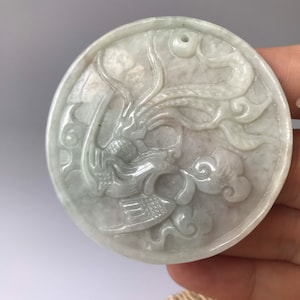 Puede incluir: Un colgante de jade verde pálido tallado con un diseño detallado de una criatura mítica, posiblemente un dragón, con nubes arremolinadas y detalles intrincados.