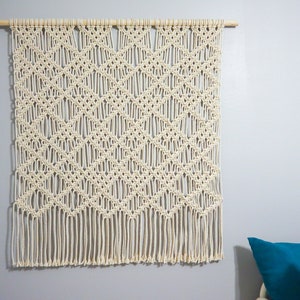 May include: A white macrame wall hanging with a diamond pattern and fringe. The hanging is made with natural cotton cord and is attached to a wooden dowel.