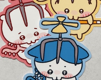 May include: Three cartoon animal stickers with propeller hats. The stickers are white with pink cheeks and are holding a stick. The stickers are on a white background. The first sticker is yellow with a gray propeller hat. The second sticker is white with a pink propeller hat. The third sticker is blue with a gray propeller hat.