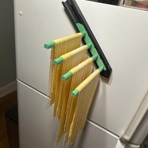 以下が含まれることがあります： 黒と緑の金属製の麺干しラックに、乾燥した麺が5本ぶら下がっています。ラックは白い冷蔵庫のドアに取り付けられています。