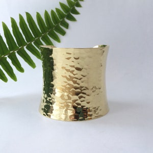 May include: A wide, gold cuff bracelet with a hammered texture. The bracelet has a curved shape and is set against a white background with a green fern leaf.
