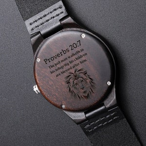 May include: Wooden wristwatch with a dark brown face and black leather strap. The watch face features the text "Proverbs 20:7" and a lion's head engraving. The watch has a silver-colored crown.