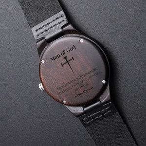 May include: A wooden wristwatch with a black leather band. The watch face is engraved with the words "Man of God" and a cross. The text below the cross reads "Follow after righteousness, godliness, faith, love, patience, meekness. 1 Timothy 6:11"