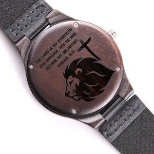 May include: Close-up of a wooden watch with a black leather strap. The watch face features engraved text and a lion's head silhouette. The text reads "THE LORD IS MY STRENGTH AND DEFENSE, AND HE HAS BECOME MY SALVATION. EXODUS 15:2."