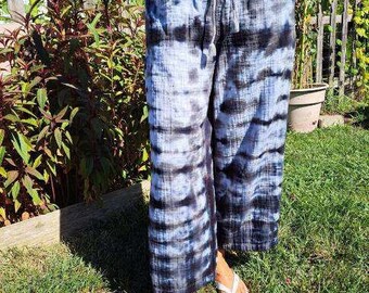 May include: Tie-dye wide-leg pants in shades of blue and black with a drawstring waist. The pants are worn with white flip-flops. The model is standing on a grassy lawn.