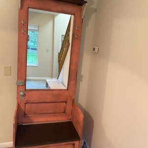 Può includere: Una panca d'ingresso in stile vintage con uno specchio e un vano portaoggetti. La panca è dipinta di un colore rosa polveroso con un piano in legno scuro e una seduta nera. Lo specchio è incorniciato in una porta di legno con uno scaffale sopra.