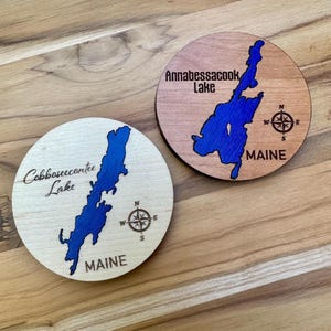 May include: Two round wooden coasters with a blue lake design. One coaster says "Cobbeseecontee Lake" and the other says "Annabessacook Lake". Both coasters have a compass rose and the word "MAINE".