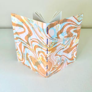 May include: Open journal with a marbled cover in shades of orange, blue, and white. The pages are white, and the binding is secured with blue thread. The journal is open, displaying the blank pages.