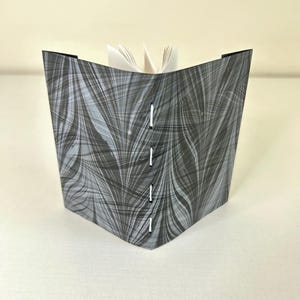 May include: A handmade journal with a marbled gray and black cover. The cover is stitched with white thread and has a unique, flowing pattern. The pages are white and visible at the top, suggesting an open book.