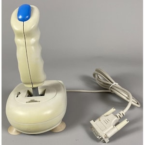 May include: A beige gaming joystick featuring a vertical handle with a blue top button, a rounded base with two small adjustment switches, and a long beige cable ending in a standard multi-pin serial connector for computer input devices.