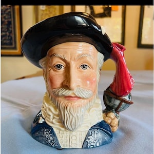 May include: A ceramic character jug depicting a historical figure with a white beard, moustache, and ruff collar. The figure wears a dark blue hat and a blue doublet with floral details. A red parrot sits on a globe attached to the handle of the jug.