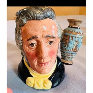 Puede incluir: Una jarra de cerámica con forma de personaje que representa a un hombre con cabello gris, vistiendo un abrigo negro y un corbatín amarillo. El asa de la jarra tiene forma de jarrón decorativo azul claro y marrón con detalles en relieve.