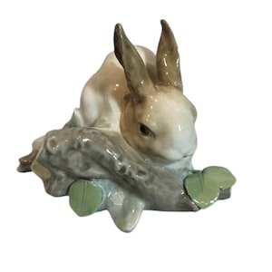 May include: A ceramic figurine of a rabbit resting on a log with green leaves. The rabbit has brown and white fur, with long ears. The log is gray and the leaves are a light green color. The figurine is on a white background.
