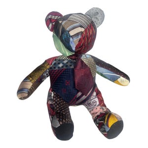 May include: A stuffed teddy bear constructed from various neckties featuring a patchwork of patterns and colors including deep red, navy blue, gold, and grey. The bear features a small necktie attached to its chest and a multi-textured fabric surface.