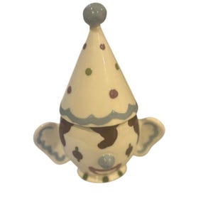May include: A ceramic clown-shaped container. The cream-coloured body has brown patches and blue ears. The conical hat is decorated with green and purple polka dots. The clown has a blue nose and a small, round, blue knob on top. A decorative item.