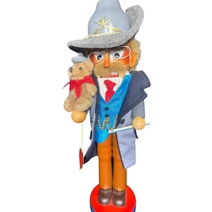 Könnte beinhalten: Eine hölzerne Nussknacker-Figur mit einem grauen Cowboyhut mit goldenen gekreuzten Schwertern, einer Drahtbrille und einem braunen Schnurrbart. Die Figur trägt eine blaue Weste, eine rote Krawatte und einen grauen Mantel.