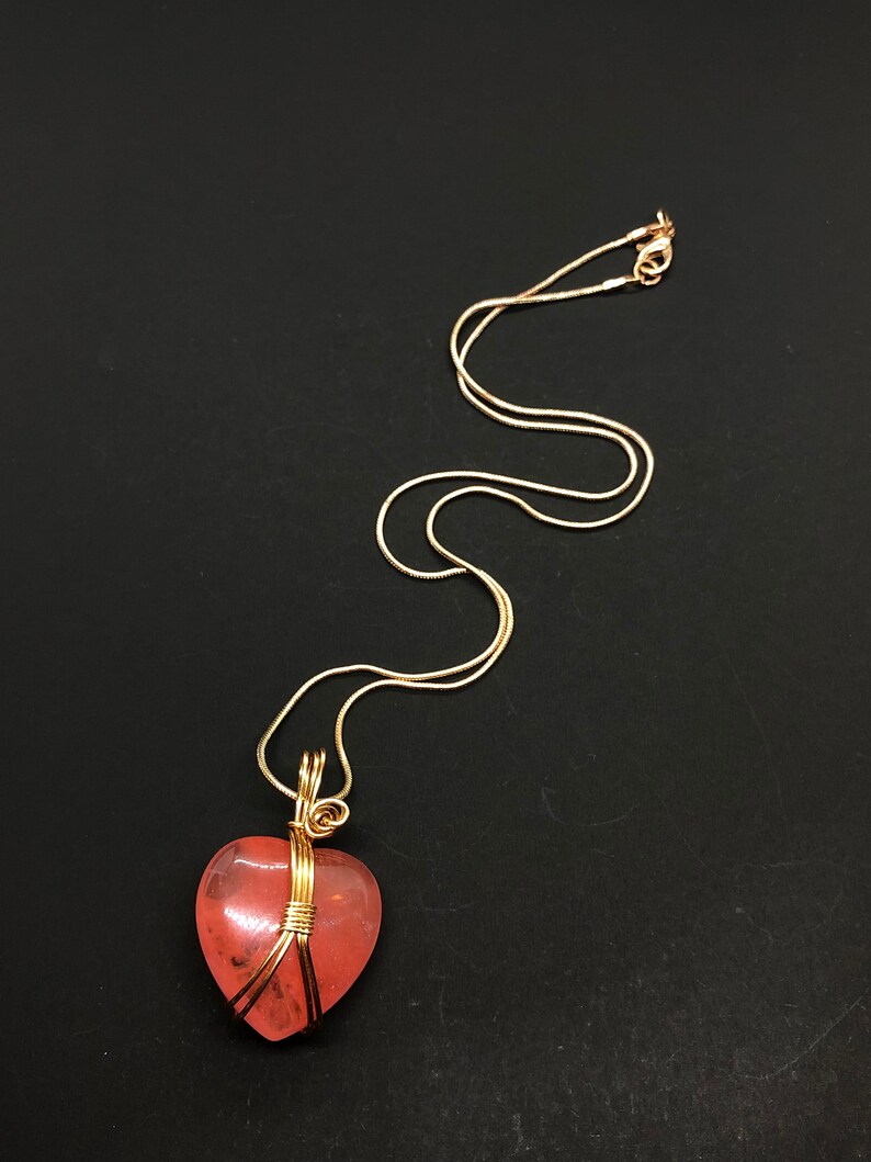 Pode incluir: Um colar de corrente de ouro com um pingente de cora&ccedil;&atilde;o rosa envolto em fio dourado.