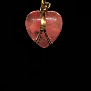 Pode incluir: Um pingente de cristal rosa em forma de cora&ccedil;&atilde;o envolto em fio dourado. O pingente est&aacute; preso a uma corrente de ouro.