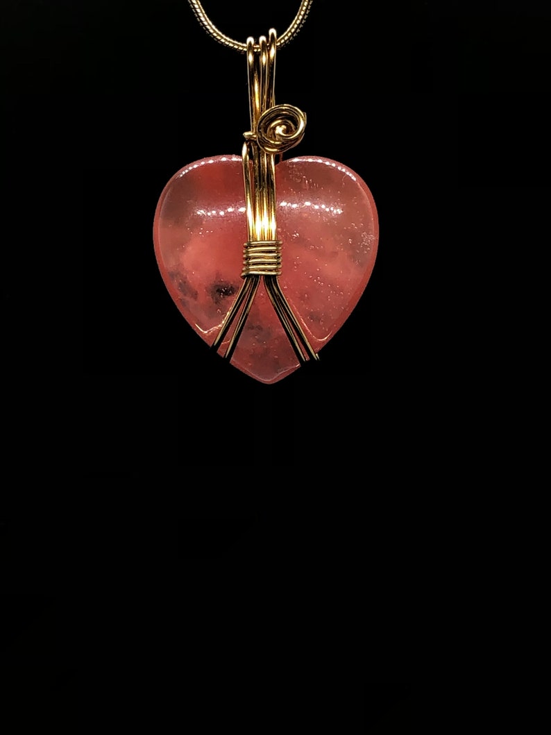 Pode incluir: Um pingente de cristal rosa em forma de cora&ccedil;&atilde;o envolto em fio dourado. O pingente est&aacute; preso a uma corrente de ouro.
