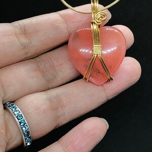 Pode incluir: Um pingente de pedra preciosa em forma de cora&ccedil;&atilde;o rosa envolto em fio de ouro, pendurado em uma corrente de ouro.