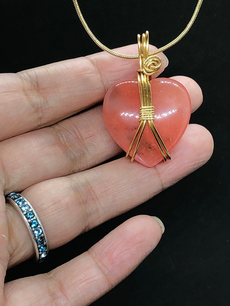 Pode incluir: Um pingente de pedra preciosa em forma de cora&ccedil;&atilde;o rosa envolto em fio de ouro, pendurado em uma corrente de ouro.