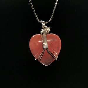 Pode incluir: Um colar de corrente de prata com um pingente de cora&ccedil;&atilde;o rosa envolto em fio de prata.