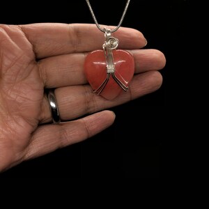 Pode incluir: Um colar de corrente de prata com um pingente de cora&ccedil;&atilde;o rosa envolto em fio de prata.