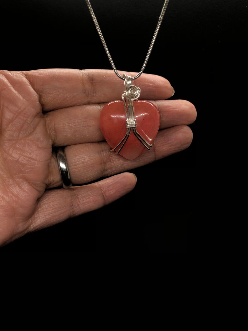 Pode incluir: Um colar de corrente de prata com um pingente de cora&ccedil;&atilde;o rosa envolto em fio de prata.