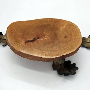 May include: A wooden serving tray with a natural, light brown finish. The tray is round with a rough, unfinished edge. Three decorative metal oak leaves are attached to the tray, two on the sides and one below.