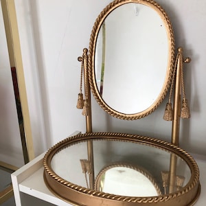May include: A gold-coloured vanity mirror with an oval-shaped mirror and a matching base. The mirror is framed with a rope-like design and has decorative tassels. The base is also oval and has a similar rope design.