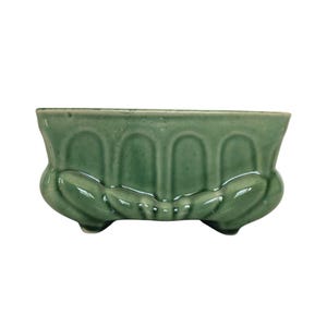 May include: A rectangular sage green ceramic planter featuring a glossy finish and a decorative molded exterior with vertical arched panels and a scalloped base design. The vessel has a wide opening and rests on small, integrated feet at the bottom.