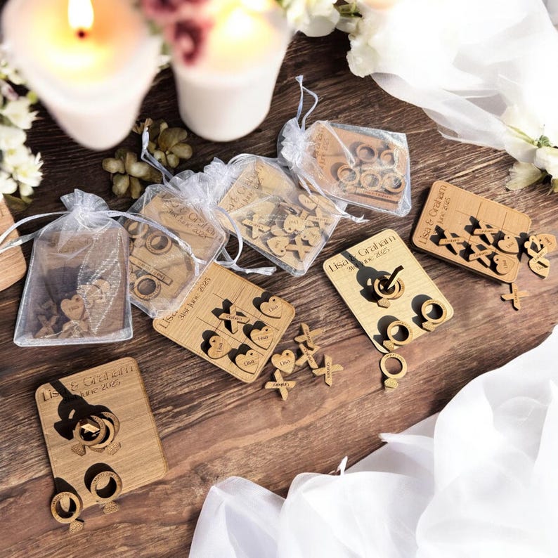 Puede incluir: Juego de boda de madera con el texto "Lisa & Graham" y la fecha "18 de junio de 2023". El juego incluye anillos, corazones y X de madera. El juego est&aacute; empaquetado en una peque&ntilde;a bolsa de organza blanca.
