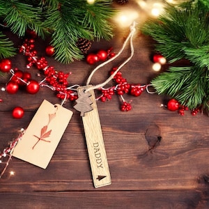 May include: A wooden Christmas tree ornament with the word "DADDY" engraved on it. The ornament is hanging from a string and is surrounded by red berries and pine branches.