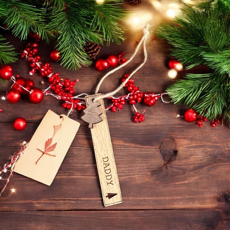 May include: A wooden Christmas tree ornament with the word "DADDY" engraved on it. The ornament is hanging from a string and is surrounded by red berries and pine branches.