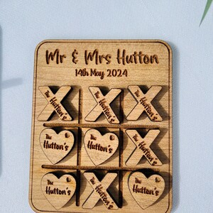 May include: A wooden tic-tac-toe game with the words "Mr & Mrs Hutton" and "14th May 2024" engraved on the board. The game pieces are heart and X shapes with the words "The Hutton's" engraved on them.