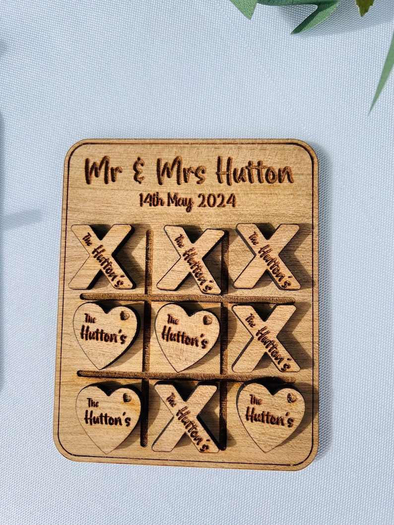 May include: A wooden tic-tac-toe game with the words "Mr & Mrs Hutton" and "14th May 2024" engraved on the board. The game pieces are heart and X shapes with the words "The Hutton's" engraved on them.