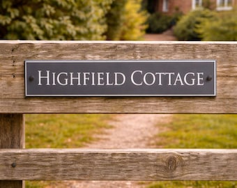 May include: A wooden fence with a rectangular black sign that reads "HIGHFIELD COTTAGE" in white lettering. The sign is mounted on the fence. A path leads to a brick building in the background.