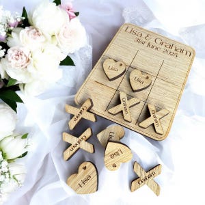 Puede incluir: Un juego de tres en raya de madera con piezas personalizadas. El tablero de juego est&aacute; hecho de madera clara y tiene los nombres "Lisa & Graham" y la fecha "31 de junio de 2025" escritos en &eacute;l. Las piezas tambi&eacute;n est&aacute;n hechas de madera y tienen los nombres "Lisa" y "Graham" escritos en ellas.