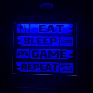 Könnte beinhalten: Ein blaues Neon-Schild mit dem Text "Eat Sleep Game Repeat" mit Symbolen für Gabel und Messer, ein Bett, einen Videospiel-Controller und ein Wiederholungssymbol.