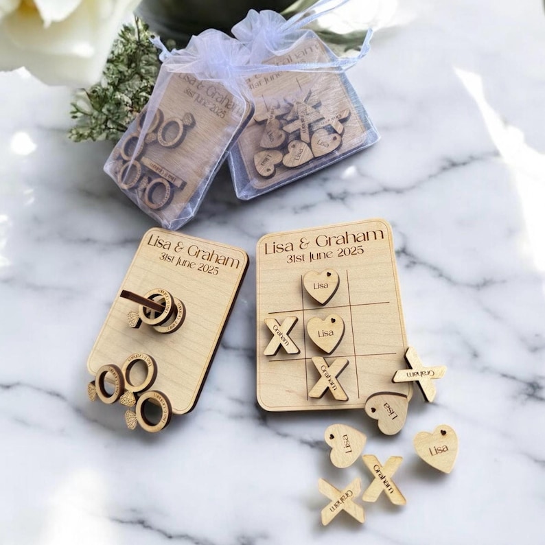 Puede incluir: Conjunto de juegos de boda de madera con dos juegos separados. Un juego es un tablero de tres en raya con piezas de madera que dicen "Lisa" y "Graham". El otro juego es un juego de lanzamiento de anillos con dos anillos y un palo de madera. Los juegos est&aacute;n personalizados con los nombres "Lisa & Graham" y la fecha "31 de junio de 2025".